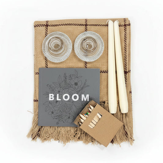 A gift bundle of decorative items including a burgundy stripe plaid blanket, taper candles, ribbed glass candle holders, and a floral coloring book with colored pencils.